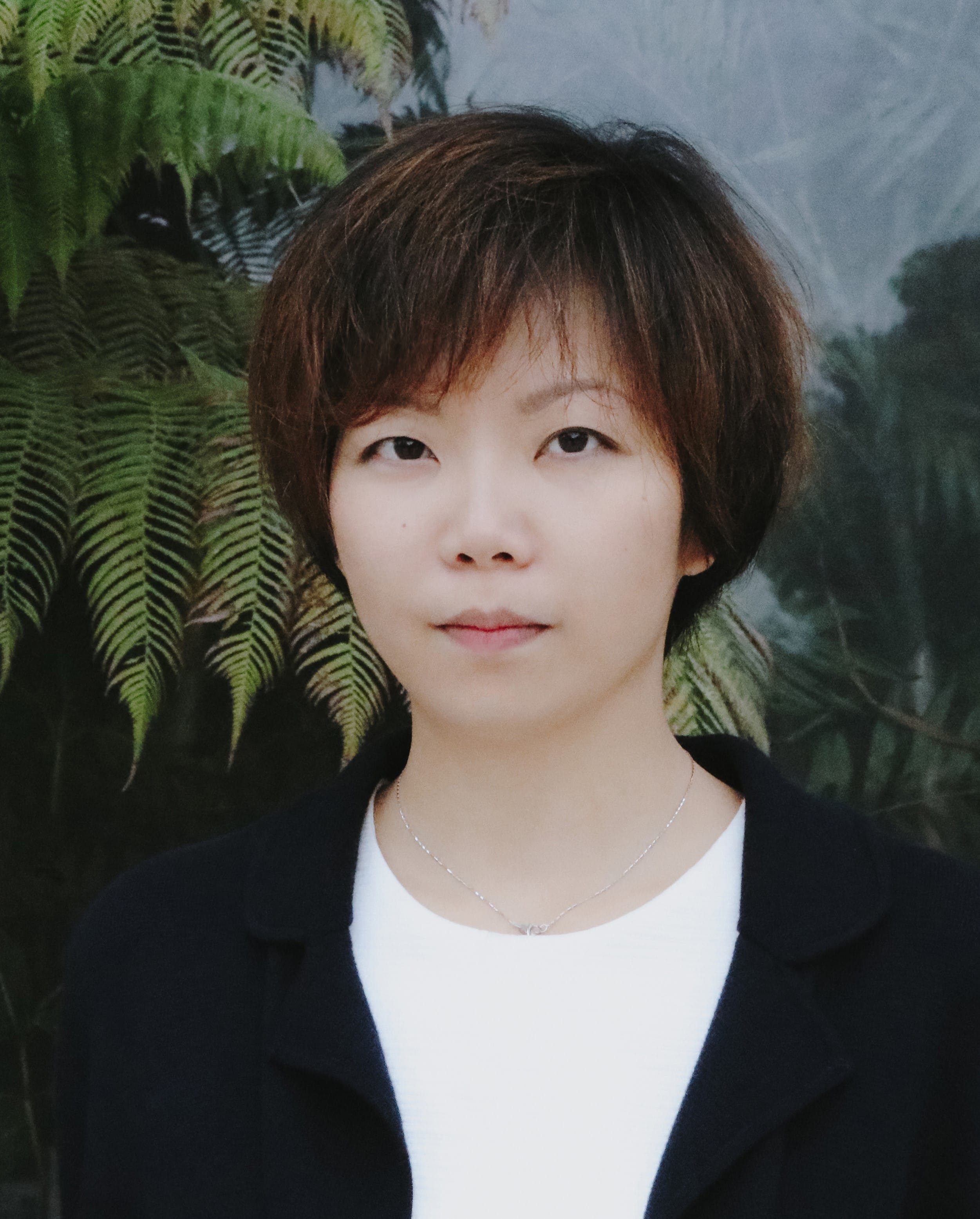 A young woman with short dark hair, wearing a white top, black jacket, and thin necklace, standing before green ferns.